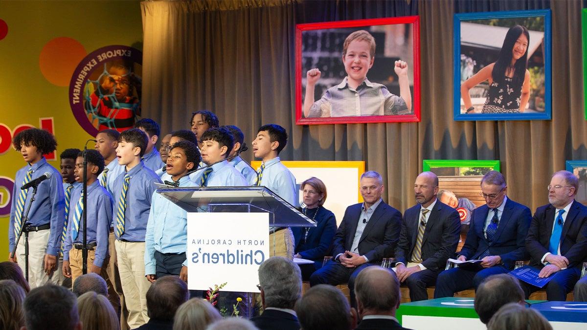 A group of of children singing on stage with a group of administrators seated behind them at an event announcing a new children's hospital in North Carolina.